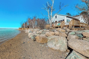 A house with a wooden deck, situated on a rocky shoreline with a clear view of the water.