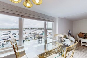 Ein modernes Wohnzimmer mit einem Glastisch, weißen Korbmöbeln und großen Fenstern mit Blick auf eine Marina.