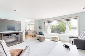 A spacious living room with a television, a fireplace, a dining area, and large windows with a view. - Charming Lakefront House with Outdoor Firepit, Washer/Dryer and Wifi (Gouldsboro)