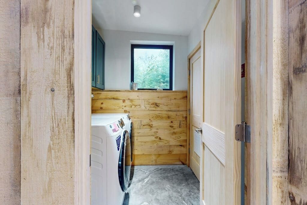 A laundry room with wooden walls, a washing machine, and a window with a view of trees.
