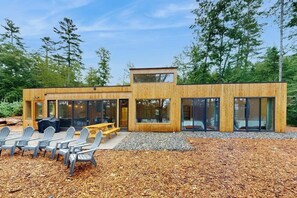 A modern wooden cabin with large glass windows, a deck with chairs, and a gravel area. - Idyllic lakeside house with outdoor sauna, patio and firepit (Hope)