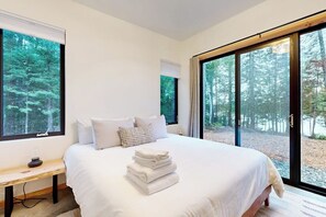 A bedroom with a large bed, a bench, and a view of a forest through large windows.
