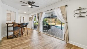 Ruime kamer met houten vloer, plafondventilator, een bar met krukken en een glazen schuifdeur naar een buitenterras met zwembad.