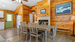 A wooden interior with a dining table, chairs, a refrigerator, a green door, and a fireplace.