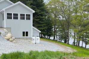 A modern house with a gravel driveway, two white water tanks, and a lake view. - Cozy Lakefront Cottage with Stunning Views, Deck, BBQ Grill, and Outdoor Firepit (Newcastle)