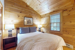 Quarto de cabana de madeira com uma cama, duas mesas de cabeceira com abajures, uma janela com persianas e um quadro na parede.