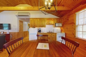 Interior de una cabaña de madera con una mesa de comedor, sillas, una zona de cocina con armarios de madera, un refrigerador y un televisor montado en la pared.
