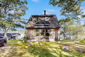 A two-story house with a chimney, large windows, and a grassy yard with rocks. - 2 Waterfront Cottages in Ocean Point with Dock & Large Yard, Dog friendly (East Boothbay)