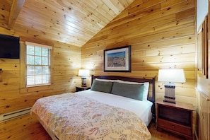 Un dormitorio de cabaña de madera con una cama, dos lámparas de noche, un cuadro enmarcado en la pared y una ventana con persianas.