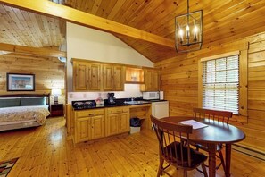 Interior de una cabaña de madera con cama, kitchenette, mesa de comedor y ventana con persianas.