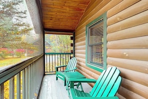 Une cabane en bois avec une véranda, des fauteuils Adirondack verts et une fenêtre avec des stores.