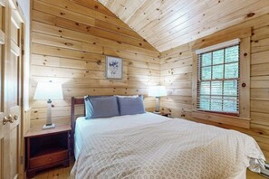 A wooden cabin bedroom with a bed, two bedside tables, a window, and a framed picture on the wall.
