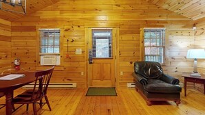 Ein Holzhütten-Interieur mit Holzboden, einem Esstisch mit Stühlen, einem Ledersofa, einem Kamin und zwei Fenstern mit Jalousien.