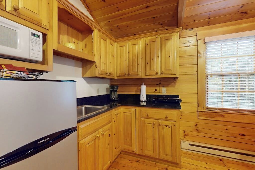 A kitchen with wooden cabinets, a microwave, a refrigerator, and a window with blinds.