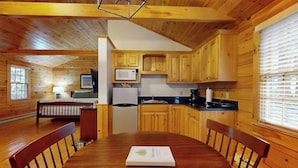 Ein Holzblockbau-Innenraum mit Esstisch, Stühlen, einer Küchenzeile inklusive Holzschränken, Kühlschrank und Mikrowelle.