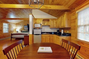 Ein Holzblockbau-Innenraum mit Esstisch, Stühlen, einer Küchenzeile inklusive Holzschränken, Kühlschrank und Mikrowelle.