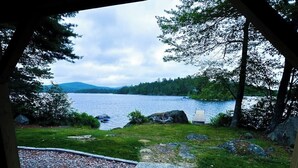 A view from a covered area overlooking a lake with a wooden deck, a stone pathway, and surrounding greenery.