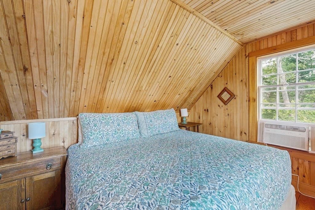 A bedroom with a sloped ceiling, wooden walls, a bed with patterned bedding, a nightstand with a lamp, and a window with a view of greenery.