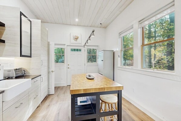 A modern kitchen with white cabinetry, a wooden island, and large windows offering a view of the forest.