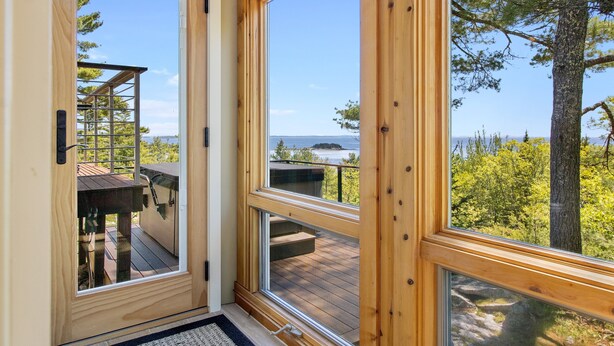 A wooden-framed window with a view of a deck and a distant body of water. - Elegant ocean view home with breathtaking views, private hot tub & large deck (Rockport)
