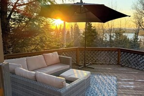 A covered outdoor seating area with a wicker sofa and a patterned rug, overlooking a forested landscape. - Scenic retreat with a spacious deck, gas fireplace, and Moosehead Lake views (Greenville)