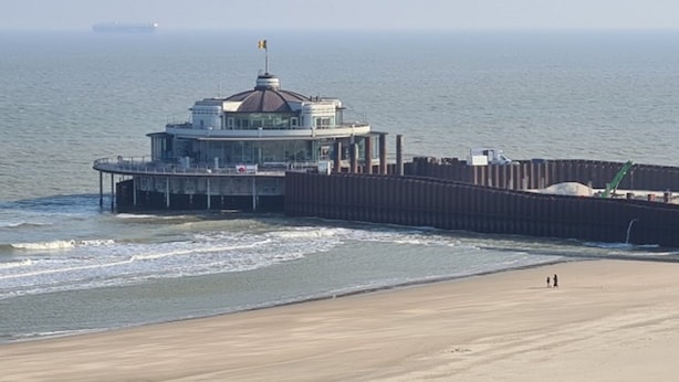 Un edificio costero con una estructura en forma de cúpula, un muelle que se adentra en el agua y una playa arenosa en primer plano.