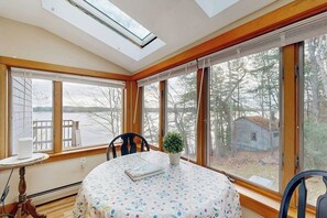 A room with a skylight, large windows, a round table with a floral tablecloth, a chair, and a small potted plant. - High View- Serene waterfront home w/ a dock, large deck & amazing views! (Harpswell)