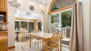 A dining area with a wooden table and chairs, a kitchen with a microwave, and a large window with curtains. - High View- Serene waterfront home w/ a dock, large deck & amazing views! (Harpswell)