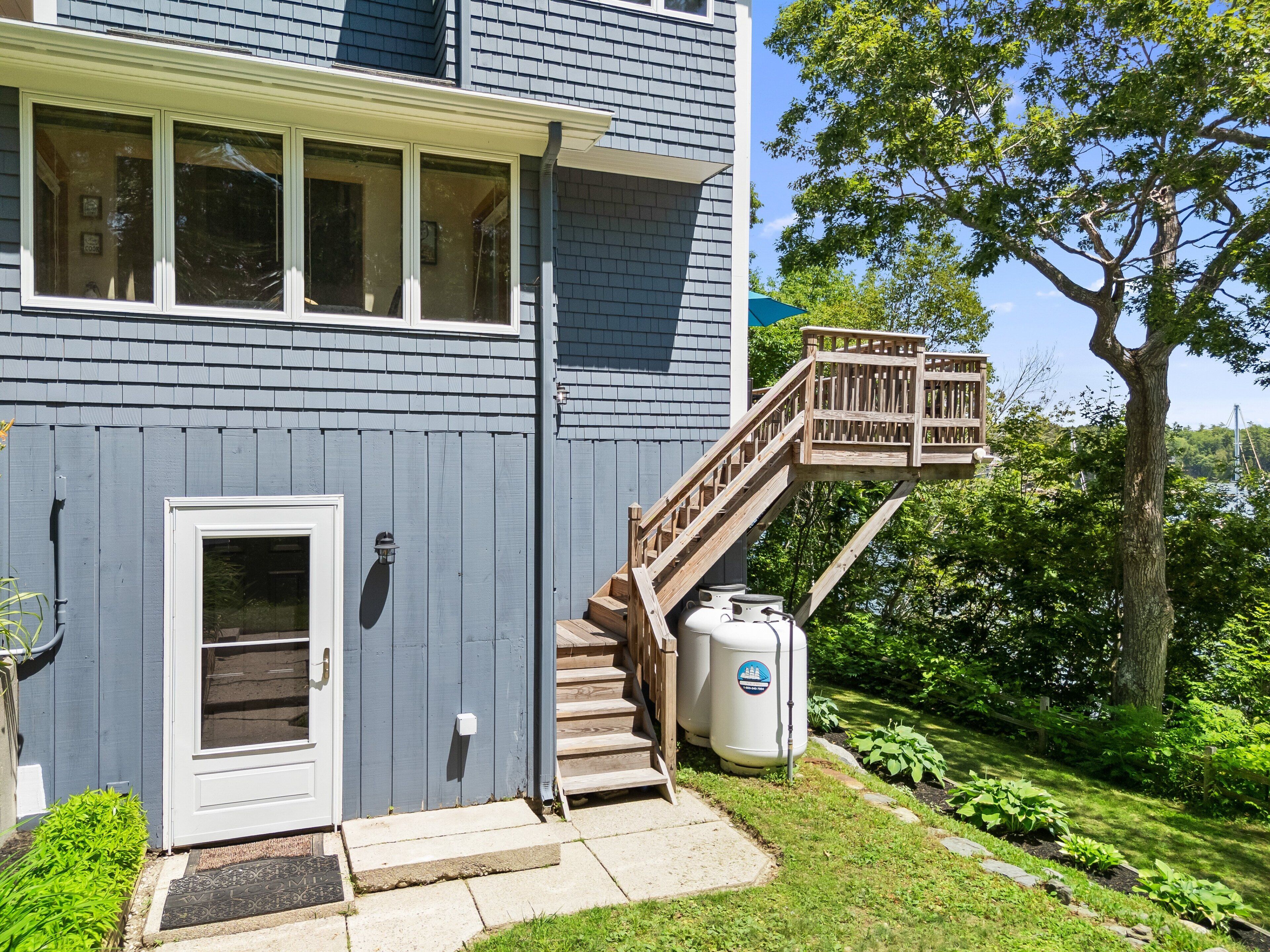 A two-story house with a front door, a wooden staircase leading to a deck, and a large tree in the background.