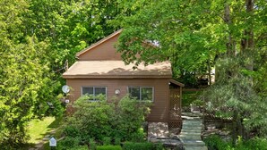 A two-story house with a sloped roof, surrounded by dense greenery and a stone pathway leading to the front door. - Ledgewood 9 - Charming Cottage near Seal Harbor Beach and hiking trails (Mount Desert)
