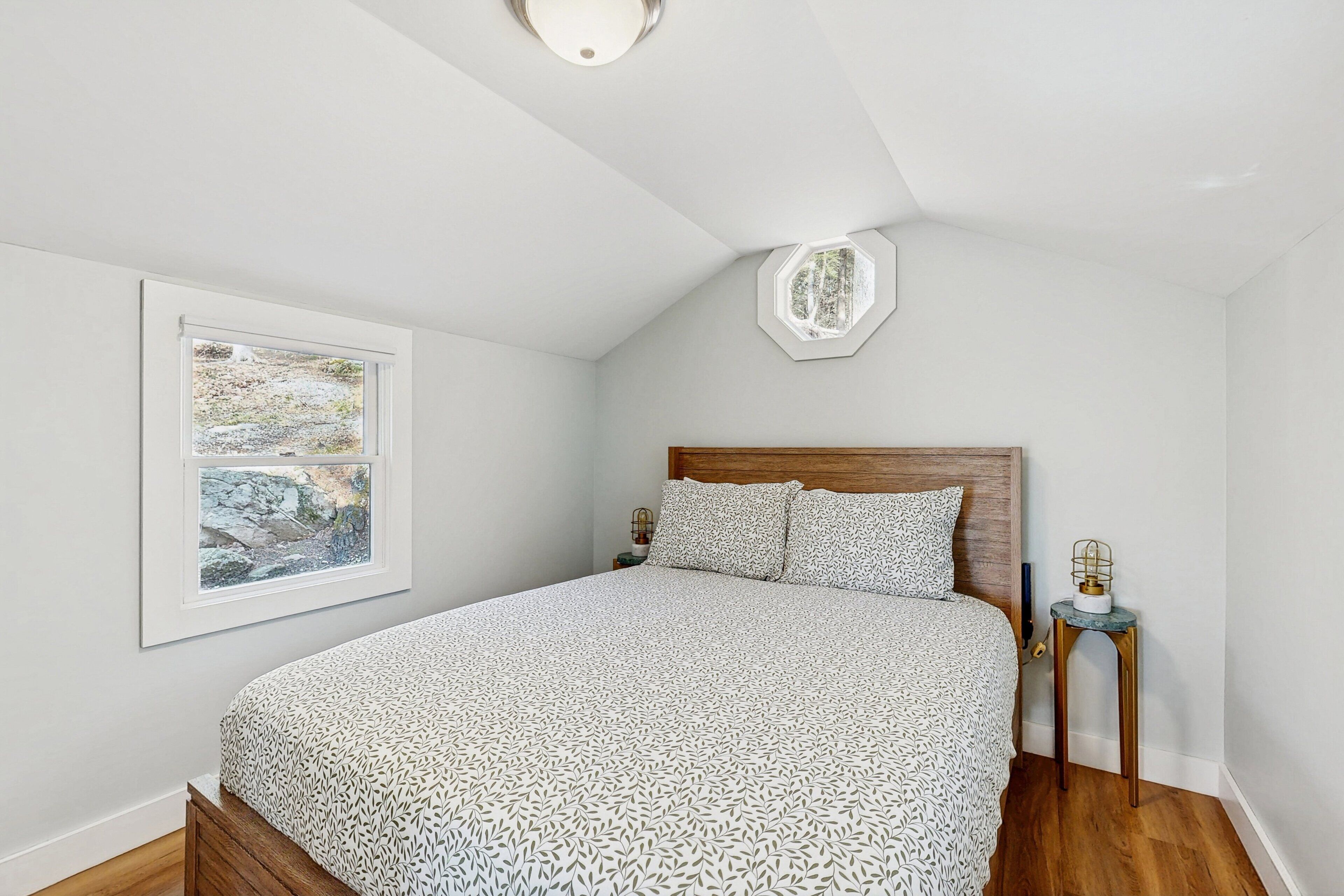 Een slaapkamer met een groot bed, een raam met uitzicht op bomen, een houten hoofdeinde en een klein tafeltje met een lamp.