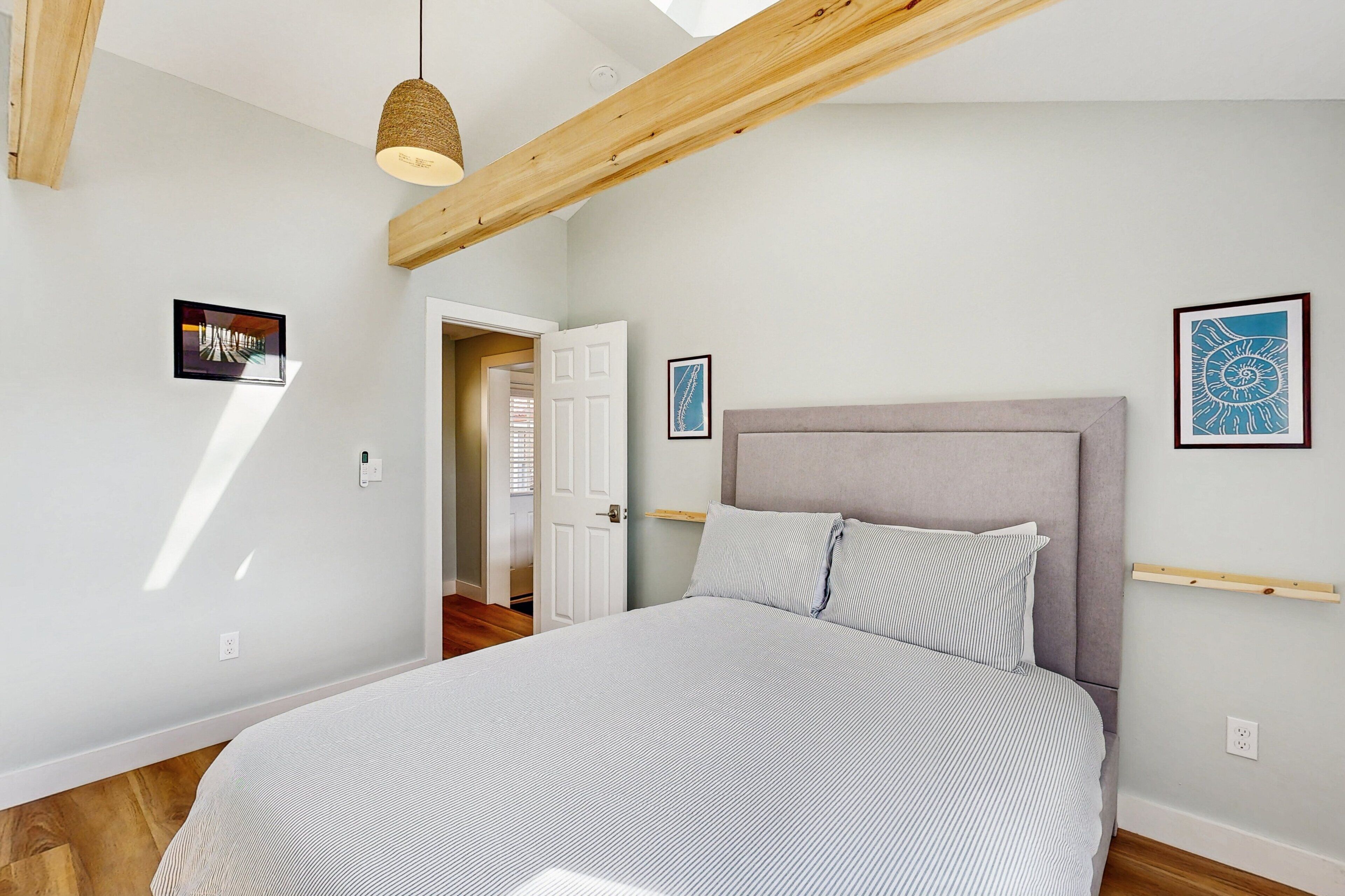 A bedroom with a bed, wooden beams, a hanging light fixture, and two framed artworks on the wall.