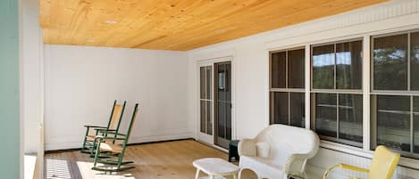 A room with a wooden ceiling, white walls, and large windows. There are two wicker chairs and a white chair.