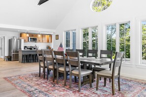 Una cocina moderna con electrodomésticos de acero inoxidable, un comedor con una mesa grande y sillas, y vistas a árboles a través de amplios ventanales.