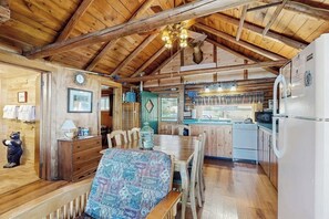 Een rustiek houten interieur met een eettafel, een bank, een keukenhoek met een spoelbak en fornuis, en een plafond met zichtbare balken.