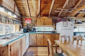 Interior rústico de una cabaña con paredes de madera, una cocina con lavaplatos, estufa y armarios, una mesa de comedor con sillas, y un letrero que dice 'YE OLD LOG CABIN'.