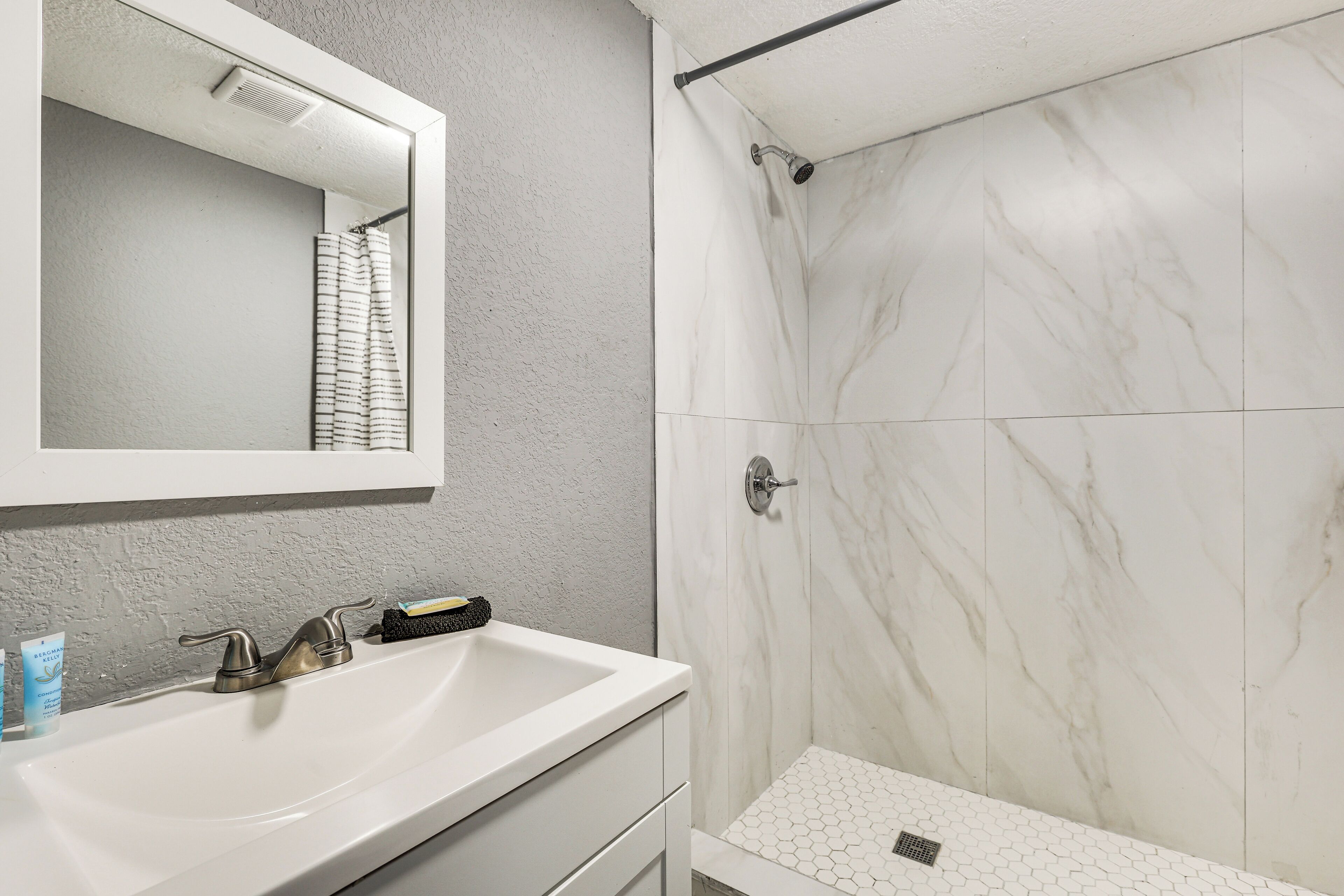 Un baño con un lavamanos blanco, un espejo, ducha con azulejos blancos y una pared con textura.