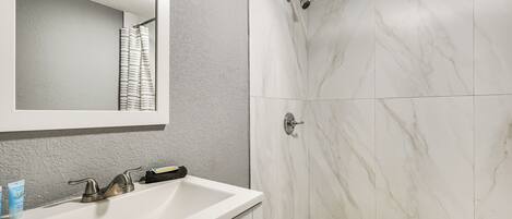 Un baño con un lavamanos blanco, un espejo, ducha con azulejos blancos y una pared con textura.