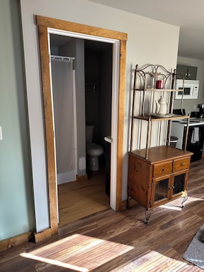 A room with a wooden floor, a white door leading to a bathroom, a kitchen area with a microwave, and a wooden cabinet with drawers. - Cozy 2-bedroom cottage in serene Dingmans Ferry with AC (Dingmans Ferry)