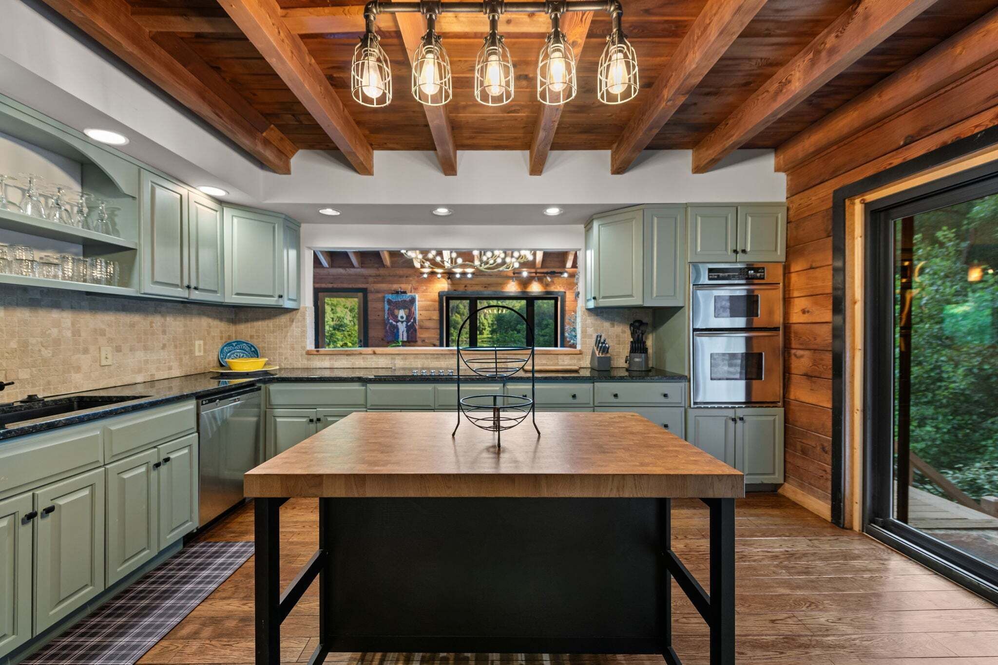 A modern kitchen with wooden beams, green cabinets, a central island, and a large window.