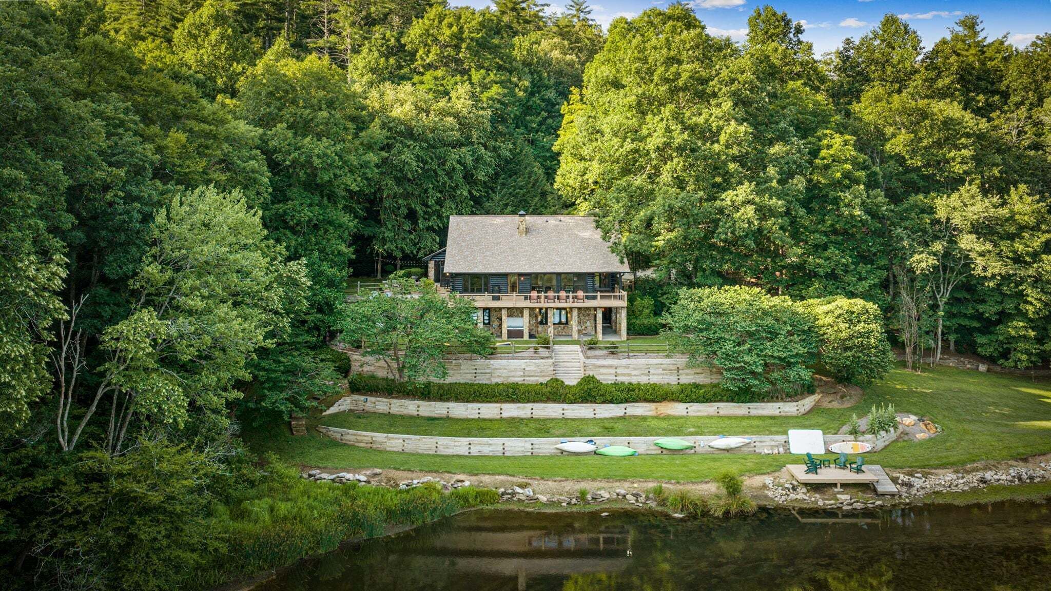 A house surrounded by dense forest, with a pond and outdoor seating area.