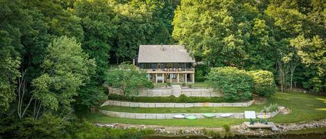 A house surrounded by dense forest, with a pond and outdoor seating area.