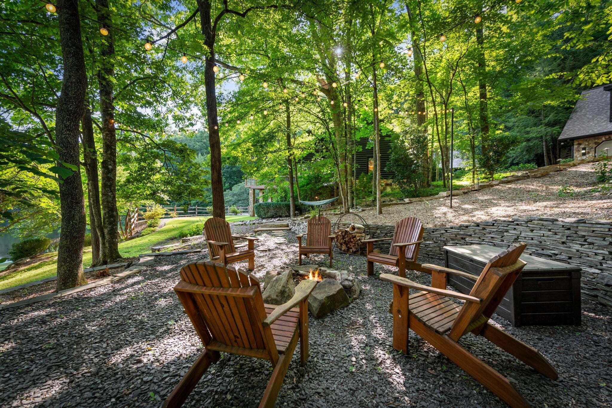 A forested outdoor area with a fire pit, wooden chairs, and a stone bench.