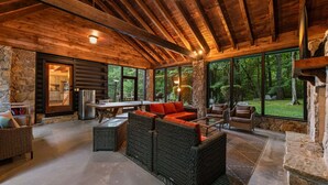 A log cabin interior with stone walls, large windows, wicker furniture, and a stone fireplace. - Swimming Bear Lodge - Lakefront | Platform Dock | Pet Friendly | Game Room (Cherry Log)