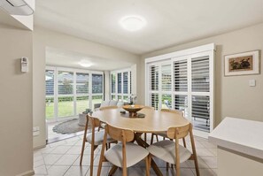 A dining area with a round table, chairs, and a view through large glass doors to a garden. - Garden Villa at Diggers (Coffs Harbour)