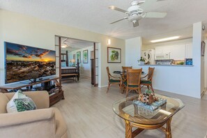 A living room with a flat-screen TV, a glass coffee table, wicker chairs, a sofa, a ceiling fan, and a kitchen area with cabinets and appliances. - Epic Sunsets. Oceanview 2 BR. Walk to Beach and Shops. (Kihei)