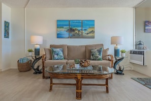 A living room with a sofa, two lamps, a glass coffee table, and artwork on the wall. - Epic Sunsets. Oceanview 2 BR. Walk to Beach and Shops. (Kihei)