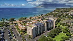 Zona costera con un edificio grande que tiene paneles solares, un estacionamiento y una playa al fondo.