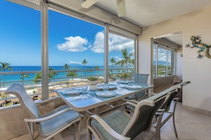 A dining area with a view of the ocean, large windows, and a table set for four. - Epic Sunsets. Oceanview 2 BR. Walk to Beach and Shops. (Kihei)