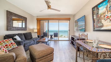 A living room with a sofa, ottoman, dining table, and chairs, a ceiling fan, and a large window with a view of the sea.